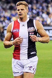 A male athlete with light hair wearing a sleeveless guernsey and shorts jogs on the grass surface of the playing arena.