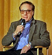 A white male in his 60s sitting in a chair while speaking into a microphone. He is wearing eyeglasses and a grey jacket over a blue buttoned down shirt. His left hand is rested on his lap.