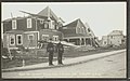 Two uniformed men stand on a sidewalk in front of a row of damaged houses.
