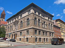 A brown stone four-story building with a partially exposed basement, wide eaves and some decorative touches on a sloping street, seen from a corner angle