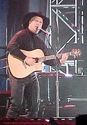 A man wearing a black cowboy hat and dark clothing, playing a guitar and singing into a microphone