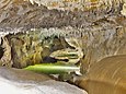 Von der Decke der sandfarbenen, beleuchteten Höhle hängen mehrere Dezimeter lange Stalagtiten. In der Mitte ist ein grünliches Wasserbecken.