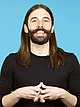 Image of Jonathan Van Ness in front of a blue background. He has long, straight brown hair and a mustache. He's smiling and is holding his hands in front of his chest.