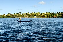 Imagem da Lagoa Mundaú. No centro da imagem há um pescador em pé em sua canoa durante a atividade da pesca. Ao fundo está a margem da laguna repleta de árvores com uma casa à direita.