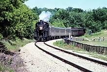 A small black steam locomotive pulling a rake of passenger coaches, with trees in the background