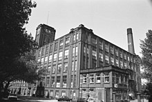 A five-storey cuboid factory composed of brick and windows. The view is from the ground at one corner of the factory. To the right, appearing from behind the factory, is a tall brick chimney. To the left are trees appearing black, and at their fringe, a square brick tower connected to the factory.