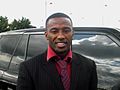 A smiling black man dressed in suit and tie stands by a large shiny car