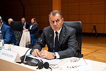 56-year-old man standing at bust length with dark jacket and tie, light blue shirt and electric blue badge holder lanyard