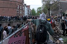 Picture taken from atop a barricade of a person in a keffiyeh sitting on top, passing a brick to someone on the outside. A group of protesters is on the other side of the canal.