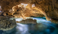 A river of blue water flowing through a brown rock cave with sun peeking through.