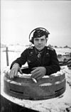 A black-and-white photo of a man protruding from the cupola of a tank wearing a black military uniform, side cap and headset.