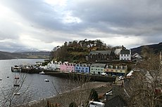 A small harbour fronted with a row of cottages painted in white, pink, green and blue with a tree-covered hillock behind them.