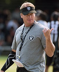 Candid chest-up photograph of Gruden on a football field wearing a grey t-shirt and a black visor