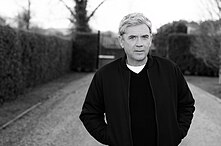 Black-and-white photograph of Mike Stock, an elderly Caucasian male with white hair, wearing a black jacket and shirt, stands in a garden