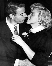 Close-up of Monroe and DiMaggio kissing; she is wearing a dark suit with a white fur-collar and he a dark suit.