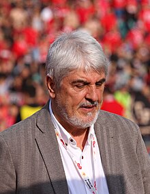 A man with white hair and beard standing in a stadium, looking toward the ground.