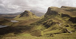 A view of green hills with steep cliffs under grey clouds. A road wends its way through the landscape.