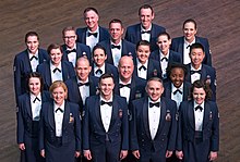This is a picture of the 19 members of the official chorus of the United States Air Force, the Singing Sergeants. They are dressed in the dark blue Air Force mess dress uniform and are standing in 5 rows on a dark brown wood floor looking and smiling up at the photographer.