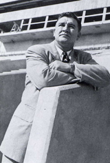 Bob Woodruff in a suit, arms folded, leaning against a wall