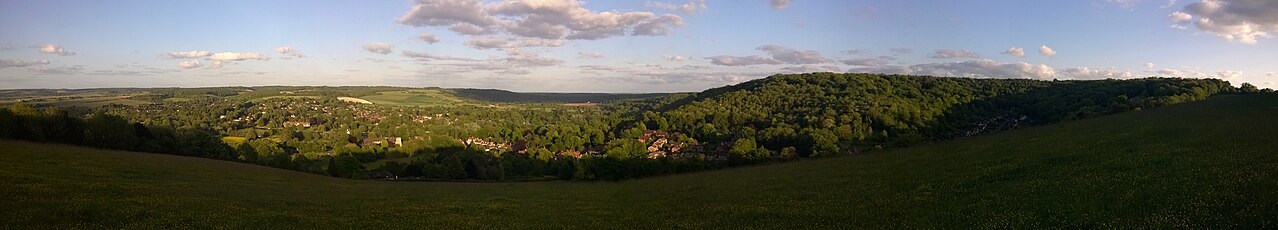 View over tree-covered hills on a sunny evening. Houses of a large settlement are visible between the trees.