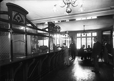 Photographie en noir et blanc de guichets devant lesquels des hommes sont attablés ou attendent leur tour.