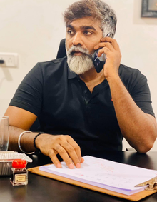 Sethupathi, holding a mobile phone next to his ear, is wearing a black shirt with a white collar, sporting a short beard and mustache, and styled in a short, spiky haircut.