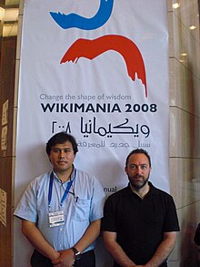 جيمي ويلز يرتدي قميص أسود وبجانبه رجل وفي الخلف لوحة عليها شعار ويكيمانيا بالعربية والإنجليزية