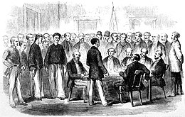 A group of seated colonial officers question a Chinese detainee as a mixed crowd of European and Chinese men watch.