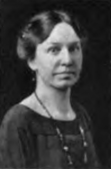 A white woman with dark hair parted center and dressed to the nape, wearing a dark top and a strand of dark beads