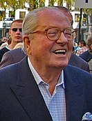 An elderly white man with receding white hair is wearing glasses. He is outdoors with various people in the background, suggesting a crowd. He is smiling, his mouth open. He is wearing a shirt with a blue jacket.