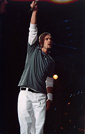 Justin Timberlake performing onstage wearing white pants and greenish jumper, with his right hand outstretched.