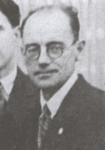 Photographie en buste et en noir et blanc d'un homme en costume qui porte des lunettes