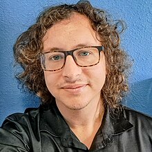 A headshot photo of a person with glasses in a black shirt against a blue background.