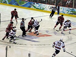 An ice hockey game. Two referees in black and white striped shirts are visible, as are four players from each team. One team is wearing red jerseys with a maple leaf on the front, and the other is wearing white jerseys with blue accents and the letters "U-S-A" on the front.