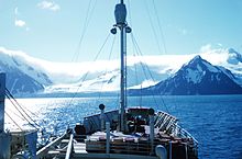 On-board view towards the bow of a small boat with a mast. Beyond the bow is a shoreline of snowy mountains.