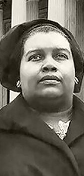 Photograph of a black woman in a dark cloche-style hat and dark coat adorned with a large shield-shaped broach, standing in front of the U.S. Supreme Court