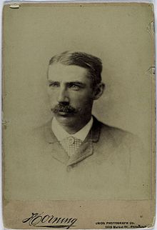 A sepia-toned image of a mustachioed man with thick, parted hair