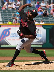 A man wearing a black baseball jersey with "Music City" written across the chest, gray pants, black socks, and a black cap in the midst of pitching a baseball from the mound