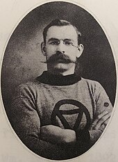 Black and white photo of a dark-haired middle-aged man wearing eyeglasses, an athletic sweater and a large moustache