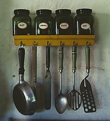 Various kitchen utensils. At top: a spice rack with jars of mint, caraway, thyme, and sage. Lower: hanging from hooks; a small pan, a meat fork, an icing spatula, a whole spoon, a slotted spoon, and a perforated spatula.