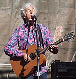 Colour photograph of Robyn Hitchcock, playing a guitar, standing and singing into a microphone with closed eyes