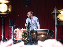 A bald, bearded man rest his hands in a timpani while a church bell is beside him