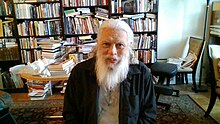 Photo couleur d'un vieil homme avec une barbe fournie et des cheveux blancs sur fond de livres de bibliothèque.