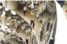 The same rotunda with the balconies covered in mirror-polished stainless steel. There are people walking along galleries, and a burly man with large beard, wearing, a black t-shirt, leans on the railing.