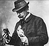 A bespectacled man wearing a hat and holding a pigeon in one hand and a small camera in the other