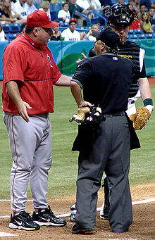 A man in a red warmup with gray pants stands arguing with a man in a black baseball jersey and dark gray pants.