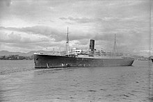 Photo noir et blanc d’un bateau à une cheminée, avec la côte proche en arrière-plan.