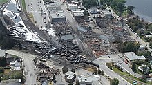 An aerial view of a small town on a lake. Over half the buildings are burnt to the ground, next to over 60 oil tanker cars derailed off a long sweeping curve. White foam covers the ground while firefighters cool the wreck.