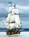 A wooden sailing vessel with numerous rope connecting from the deck to several large white sails above. An American flag drapes down from a pole at the back of the ship and a Washington state flag is on the tallest mast. The ship is in dark blue water, and in the background are a shoreline, treeline and cloudy sky.