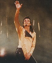 In the center for the photo, a light skinned male with black hair wearing a gold shirt and black pants, has his right hand to his crotch and his left hand held up high.